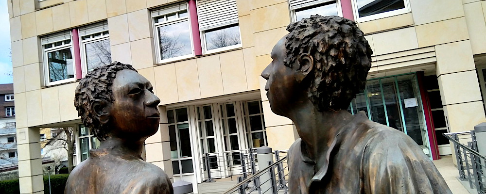 Bronzetatue vor dem Landratsamt in Freiburg. Zwei lebensgroße Personen laufen aneinander vorbei und schauen sich an..